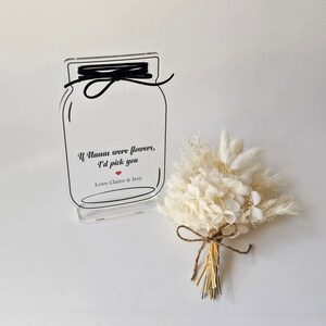 May include: A clear acrylic sign with a mason jar design and the text "If Nanas were flowers, I'd pick you" with a red heart and "Love Claire & Issy". A small bouquet of dried white flowers is in front of the sign.