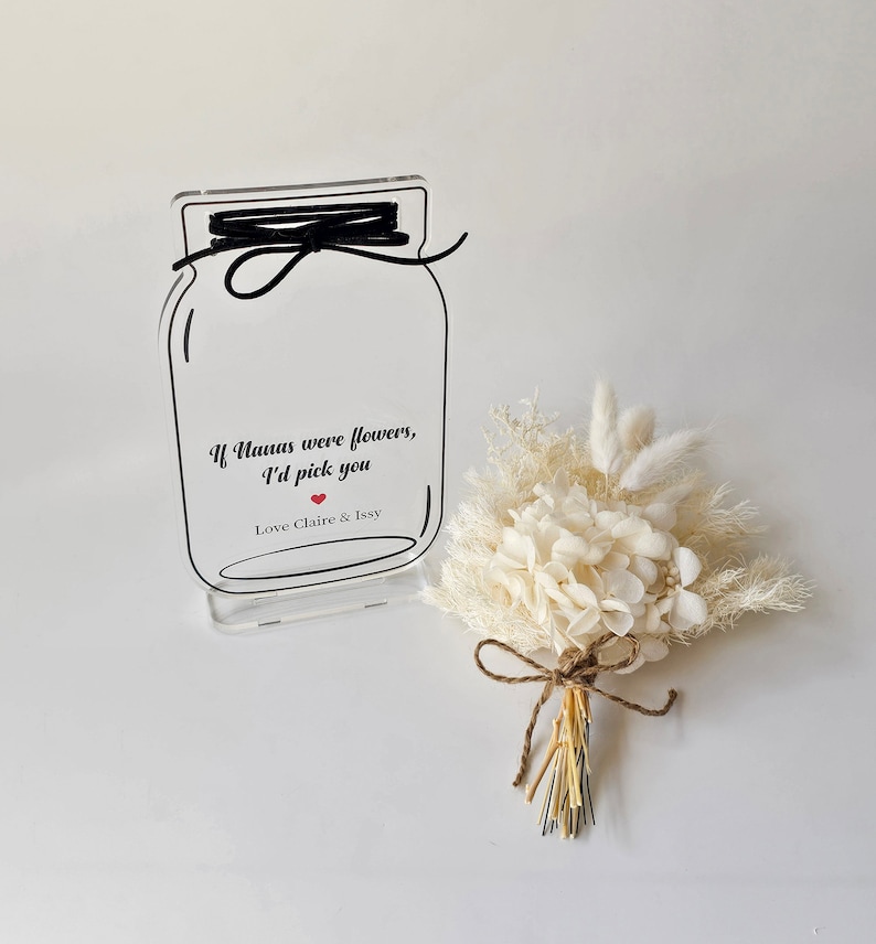 May include: A clear acrylic sign with a mason jar design and the text "If Nanas were flowers, I'd pick you" with a red heart and "Love Claire & Issy". A small bouquet of dried white flowers is in front of the sign.