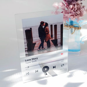 May include: Clear acrylic photo display featuring a couple kissing on a pier. The display includes the text "Love Story" by Taylor Swift, with a play button and a music timeline. A vase of dried flowers is in the background.