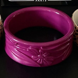 Könnte beinhalten: Ein violettes Plastik-Armreif mit Blumenmuster. Der Armreif ist mit einer Blume und Blättern geprägt. Der Armreif ist von Marion Godart Paris.