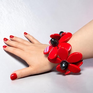 Può includere: Un bracciale caratterizzato da molteplici grandi componenti lucidi a forma di fiore rosso con centri scuri e arrotondati. I petali sono lisci e arrotondati, disposti a grappolo per formare un audace design floreale sul polso.