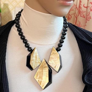 Può includere: Una collana statement nera e dorata con tre grandi pendenti geometrici sfaccettati. La collana è realizzata con perline nere e presenta un materiale dorato iridescente simile alla madreperla.