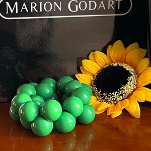 May include: A cluster of bright green round balls arranged in a circular shape, resembling a flower bud. A yellow sunflower with a dark brown center is in the background.
