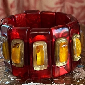 May include: A bold, red and gold bracelet. The bracelet is made of rectangular, translucent red segments, each with a gold-colored frame and a yellow-orange center. The bracelet is resting on a decorative surface.