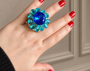 May include: A statement ring featuring a large, round, faceted royal blue center stone surrounded by a halo of smaller, pear-shaped, light turquoise blue crystals arranged in a floral pattern, set on a metallic base with a polished finish.