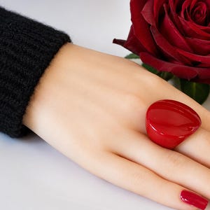 Può includere: Un grande anello rosso liscio e lucido con una forma organica arrotondata. L'anello presenta una finitura rossa solida e vibrante che cattura la luce, creando un aspetto lucido. È indossato su un dito, completato da uno smalto rosso abbinato.