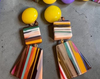 May include: A pair of drop earrings featuring a yellow spherical stud, a square block with horizontal stripes of yellow, green, white, and brown, and a trapezoidal pendant with vertical multicoloured stripes in yellow, brown, white, and teal.
