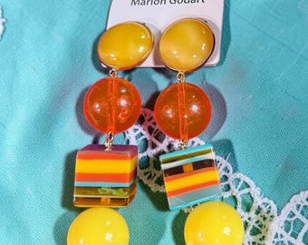 May include: Marion Godart drop earrings featuring a vertical arrangement of yellow and orange spheres, and a square bead with horizontal stripes in yellow, orange, and blue. The earrings are attached to a white display card with the brand name.
