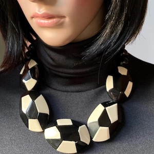 May include: A statement necklace featuring large, chunky, geometric beads in a high-contrast black and cream color palette. The beads are faceted with irregular, angular shapes, creating a bold, segmented pattern that rests against a black turtleneck.