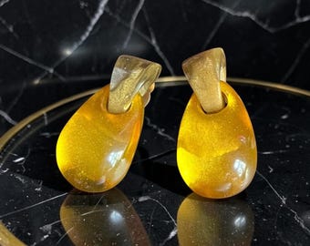 Puede incluir: Un par de pendientes colgantes con forma de gota hechos de resina translúcida de color amarillo dorado, suspendidos de piezas superiores de resina sólida con destellos dorados. Los pendientes tienen un acabado liso y pulido muy brillante.