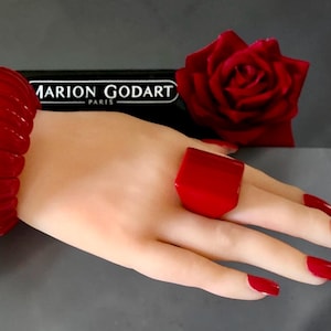 May include: A bold, square-shaped red ring with a smooth, glossy finish. The ring is worn on a finger, accompanied by a matching red bangle bracelet on the wrist. In the background, a black sign displays the text MARION GODART PARIS.