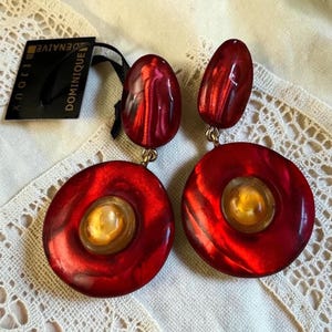 May include: A pair of drop earrings featuring a glossy, deep red, marbled resin oval top piece connected to a larger, circular red resin disc with a central, translucent amber-colored cabochon. A black tag labeled DOMINIQUE BIJOUX is attached.