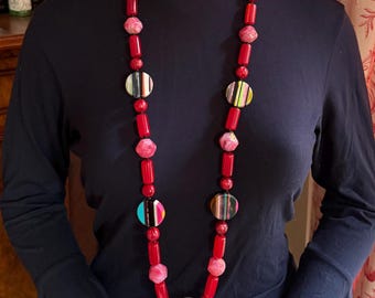 May include: A long, red beaded necklace with colorful striped and pink accents. The necklace is displayed on a mannequin wearing a black turtleneck and hat. The necklace features a variety of shapes and sizes of beads.