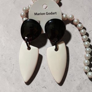Puede incluir: Pendientes colgantes Marion Godart con una pieza superior circular negra brillante conectada a un colgante liso en forma de escudo de color crema. Los pendientes están sujetos a una tarjeta blanca con el nombre de la marca en negro.