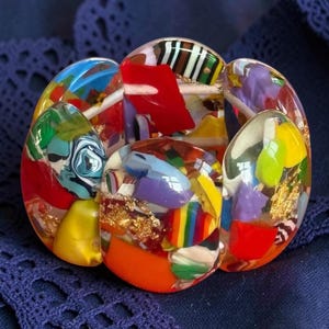 Könnte beinhalten: Ein klobiges Armband aus klaren Harzsegmenten, die mit einer lebendigen Auswahl an bunten Fragmenten eingebettet sind, darunter rote, gelbe, orange, blaue, grüne und violette Teile, akzentuiert mit Flocken aus goldener Metallfolie.
