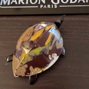 Puede incluir: Un broche de tortuga marrón, amarillo y verde con detalles negros. El broche está hecho de resina y tiene un acabado brillante. El broche está diseñado por Marion Godart Paris.