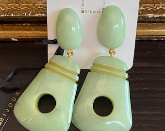 May include: A pair of green and gold statement earrings with a geometric design. The earrings feature a large oval-shaped top piece and a smaller, rectangular bottom piece with a circular cutout. The earrings are on a white card with the text "Bijoux Dominique Denaive" printed on it.