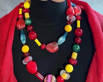 May include: A multi-strand necklace featuring an assortment of beads in various shapes, sizes, and colors including bright yellow, deep red, translucent teal, and marbled green, finished with a central circular pendant with vertical stripes.
