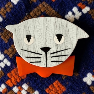 May include: A cat-shaped brooch featuring a white face with a black crackle pattern, black triangular eyes, a black nose, and black whiskers. The cat is wearing a solid, bright orange bow tie positioned directly beneath its chin.