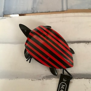 Puede incluir: Un broche de tortuga con rayas rojas y negras con una etiqueta negra que dice "MARION GODART".
