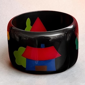 Peut inclure: Bracelet jonc noir avec des formes géométriques colorées. Le bracelet présente une maison avec un toit rouge et des murs bleus, des arbres verts et d'autres formes colorées. Un accessoire audacieux et artistique.