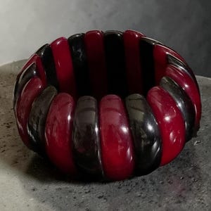 May include: A chunky stretch bracelet composed of alternating vertical segments in deep glossy black and translucent dark red. The rounded, smooth-surfaced pieces are linked together to form a continuous circular band with a polished finish.