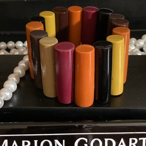 May include: A stretch bracelet composed of cylindrical resin segments in a multi-coloured palette of orange, yellow, brown, black, and magenta. The bracelet is displayed with a black rectangular plaque featuring the white text MARION GODART PARIS.