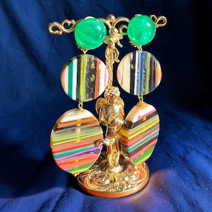 Op de afbeelding: Een paar hangende oorbellen met felgroene ronde knopjes en twee gestapelde ronde hangers. De hangers tonen verticale en horizontale strepen in diverse kleuren, waaronder zwart, wit, geel en oranje, opgehangen aan een gouden standaard.