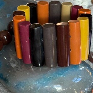 May include: A stretch bracelet composed of vertical, cylindrical beads in a variety of colours including orange, yellow, black, dark brown, maroon, and tan. The beads are arranged in a circular pattern, creating a multi-coloured, smooth, and glossy accessory.