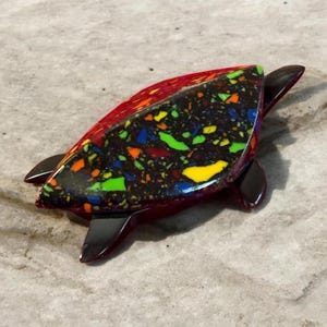 May include: A decorative turtle figurine with a red shell and black flippers. The shell features a mosaic design with colorful specks of yellow, green, blue, and orange on a dark background. The figurine is set against a neutral background.