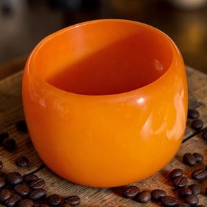 May include: A smooth, rounded, bright orange cup with a wide, open top and a thick, solid wall construction. The cup features a glossy, uniform finish throughout its exterior surface, presenting a simple, handleless, and cylindrical shape.