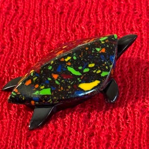 Puede incluir: Figura decorativa de tortuga con cuerpo negro y caparazón moteado. El caparazón presenta motas amarillas, verdes, naranjas, azules y rojas. La figura está sobre un fondo rojo texturizado.