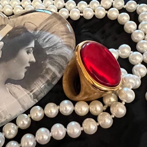 Könnte beinhalten: Ein goldener Ring mit einem großen, ovalen, roten Edelstein. Der Ring liegt auf einer weißen Perlenkette.