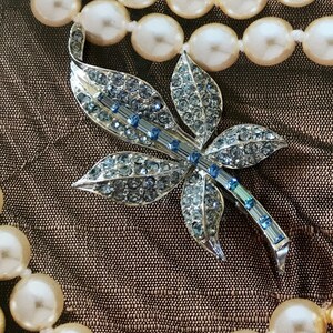 May include: A silver and blue rhinestone brooch in the shape of a leaf. The brooch is sitting on a brown fabric background with a pearl necklace.