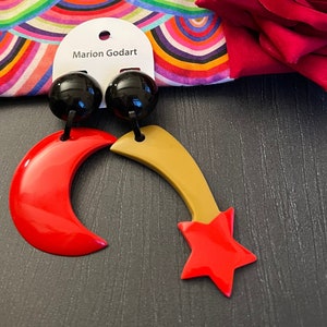 Puede incluir: Pendientes Marion Godart con tachuelas de bola negras, medias lunas rojas y diseños de estrellas fugaces doradas. Los pendientes se muestran sobre un fondo estampado colorido y una rosa roja.