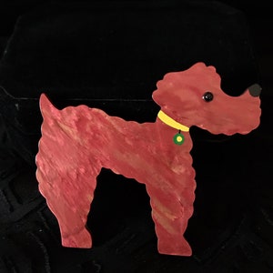May include: A red and gold-toned dog-shaped brooch. The brooch has a yellow collar with a green and yellow tag. The dog has black eyes and a black nose. The brooch is made of a material with a marbled effect.