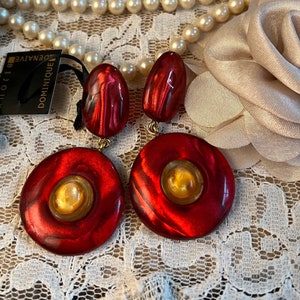 Puede incluir: Un par de pendientes de declaración redondos de color rojo y dorado con un diseño de remolino. Los pendientes están sobre un fondo de encaje blanco. La etiqueta dice "DOMINIQUE DENATIVE BIJOUX".