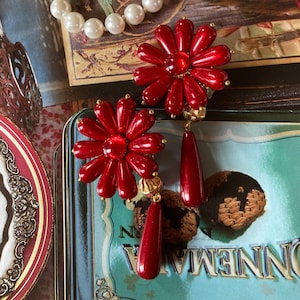 Peut inclure: Une paire de boucles d'oreilles en forme de fleur rouge avec des accents dorés et des breloques en forme de larme rouge.