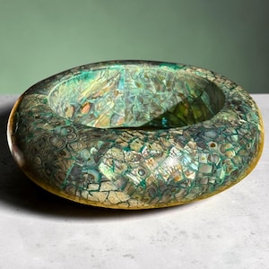 Può includere: Una ciotola circolare caratterizzata da una superficie a mosaico composta da frammenti di conchiglia di abalone iridescente in tonalità verde scuro, verde acqua e marrone. Le superfici esterne e interne presentano una finitura riflettente e multicolore.