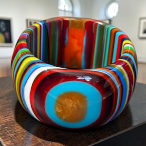 May include: A thick, circular bangle bracelet made of multi-coloured glass. The surface features vertical stripes in shades of red, orange, yellow, green, blue, and white. A prominent circular turquoise accent with a central amber-coloured spot is visible.