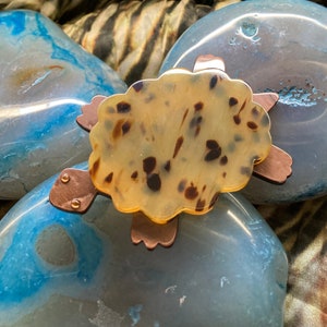 Puede incluir: Un broche de tortuga con un patrón de caparazón de tortuga marrón y blanco y detalles en cobre. El broche está sobre una piedra de ágata azul y blanca.
