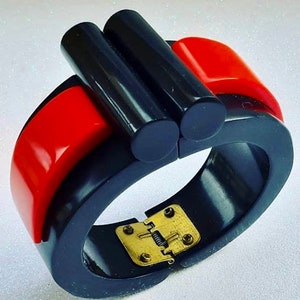 Può includere: Un braccialetto vintage audace, nero e rosso. Il braccialetto a cerniera presenta una chiusura rettangolare color oro e due accenti cilindrici neri. Le sezioni rosse hanno una finitura lucida, in contrasto con il nero opaco.