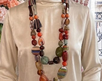 Pode incluir: Um manequim usando um chapéu cloche preto e um top de gola alta creme. Um colar longo de contas multicoloridas com pedras laranja, marrom e verde é drapeado ao redor do pescoço. Um buquê de flores cor-de-rosa está ao fundo.