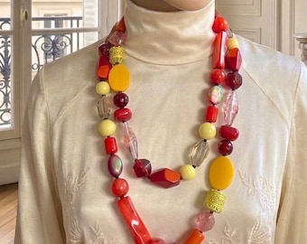 Pode incluir: Um manequim usando uma boina laranja e um colar de várias voltas. O colar apresenta uma variedade de contas nas cores vermelha, amarela e transparente. O manequim também está usando uma gola alta creme.
