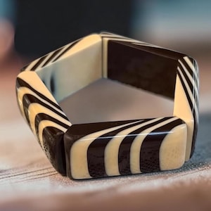 May include: A pentagonal bangle bracelet featuring a high-gloss finish with alternating segments of solid dark brown and cream-colored resin. The design displays a repeating pattern of curved, flowing stripes that wrap around each of the five flat sides.