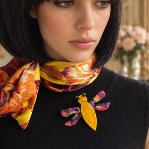 May include: A dragonfly-shaped brooch featuring a bright yellow body and two pairs of patterned wings with shades of purple, pink, and orange. The insect-themed accessory has two small black circular eyes and is pinned to a black sleeveless top.