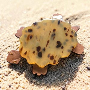 May include: A decorative turtle-shaped hair clip. The clip features a light brown shell with dark brown spots and brown legs and head. The clip is resting on a sandy surface.