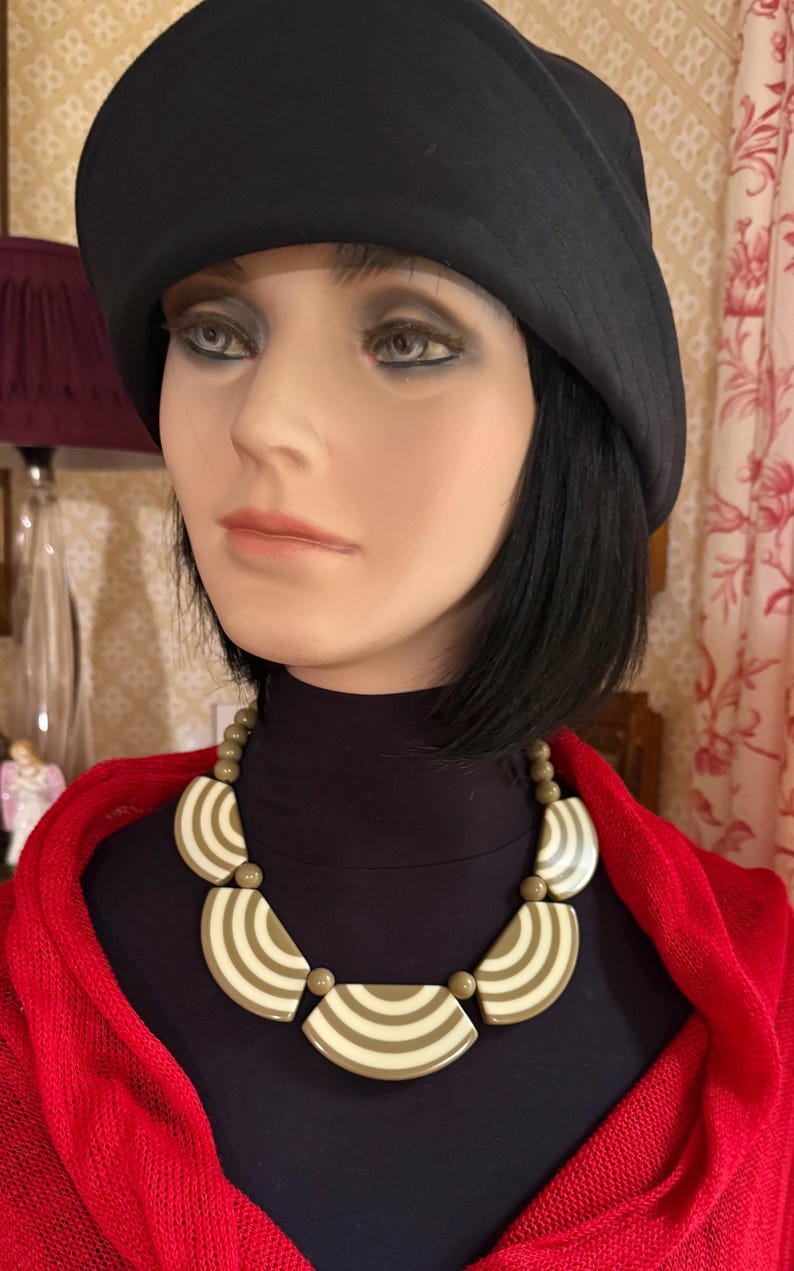 May include: A striking necklace with olive green and cream-coloured semi-circular pendants, each with striped detailing. The necklace is worn over a black turtleneck and a red shawl. A black hat is also worn.