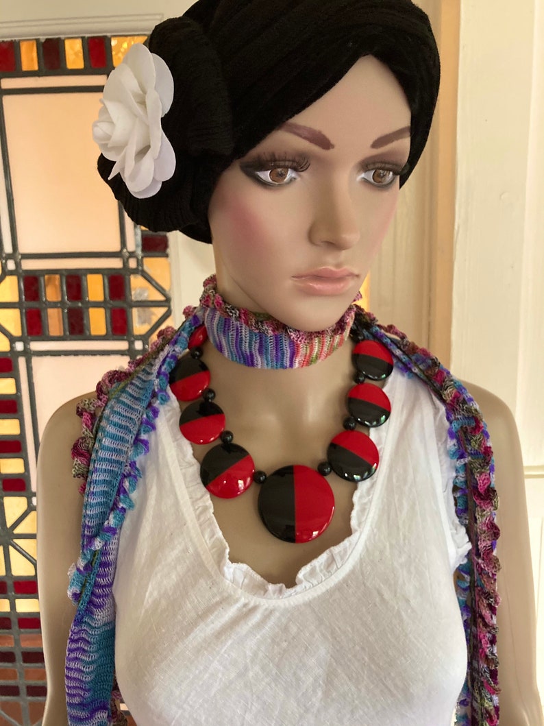 May include: A mannequin wearing a white tank top with a black and red statement necklace. The necklace is made of large, round, black and red plastic beads. The mannequin is also wearing a colorful scarf around her neck.