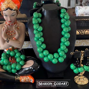 May include: A set of green and black beaded jewelry, including a necklace, bracelet, and earrings. The necklace and bracelet are made of large, round beads, while the earrings are smaller and have a dangling design. The jewelry is displayed on a black mannequin and a gold stand.